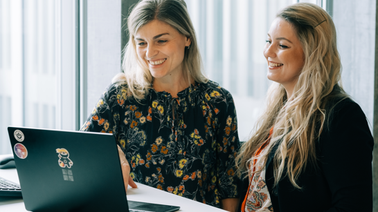 Zwei blonde Frauen vor einem schwarzen Surface Laptop lachend am reinschauen